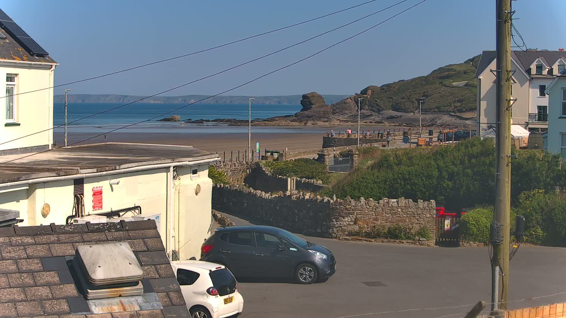 A sunny seaside view shows a sandy beach, calm ocean, and a rocky outcrop, with buildings, cars, and a low stone wall in the foreground.