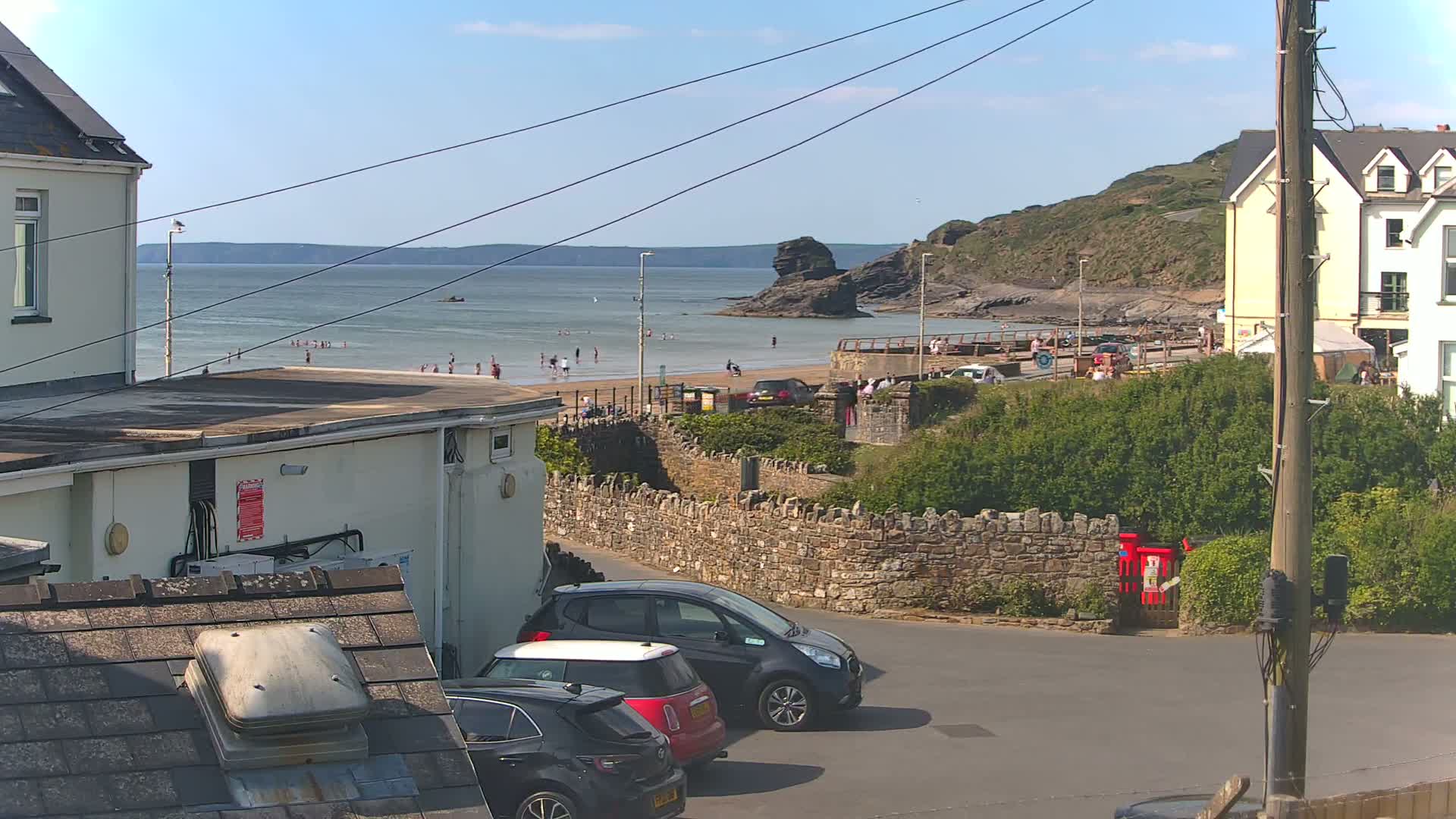 A sunny coastal scene shows a sandy beach with people swimming and sunbathing, overlooked by buildings and parked cars, with a stone wall separating the road from the beach area.