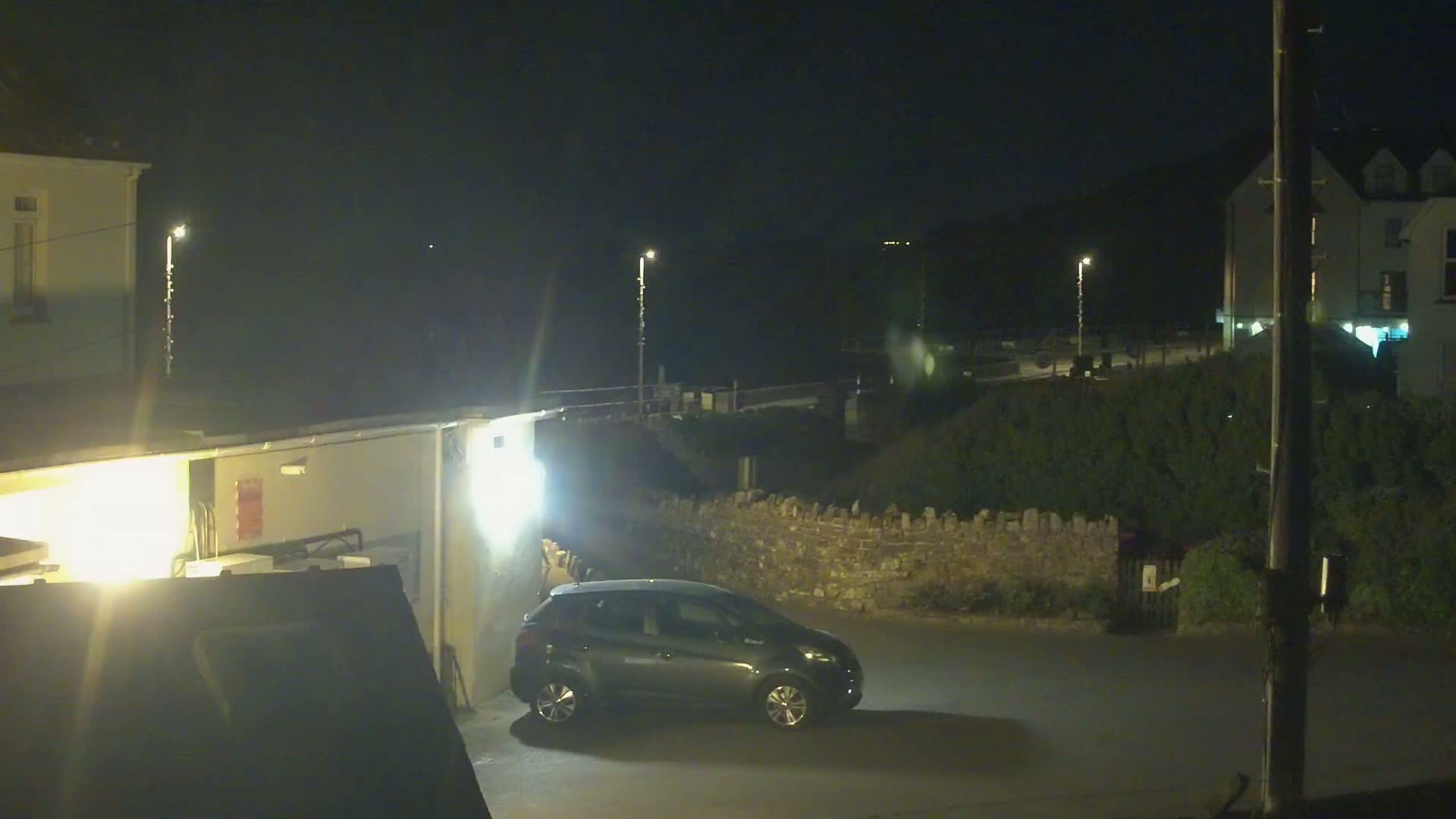 A dark-colored car is parked in a dimly lit area at night near a low stone wall and buildings.