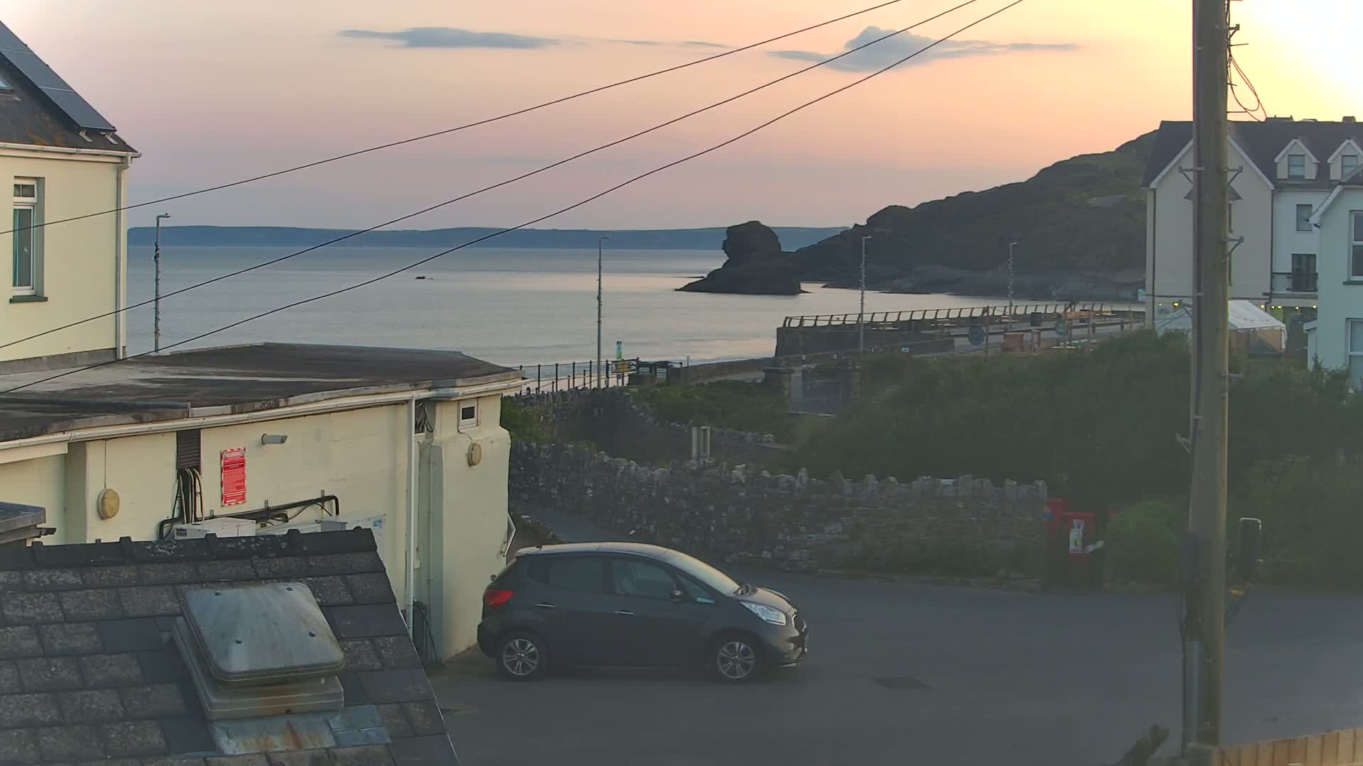A dark-colored car is parked near a seaside town at sunset, with calm water and a rocky headland visible in the distance.