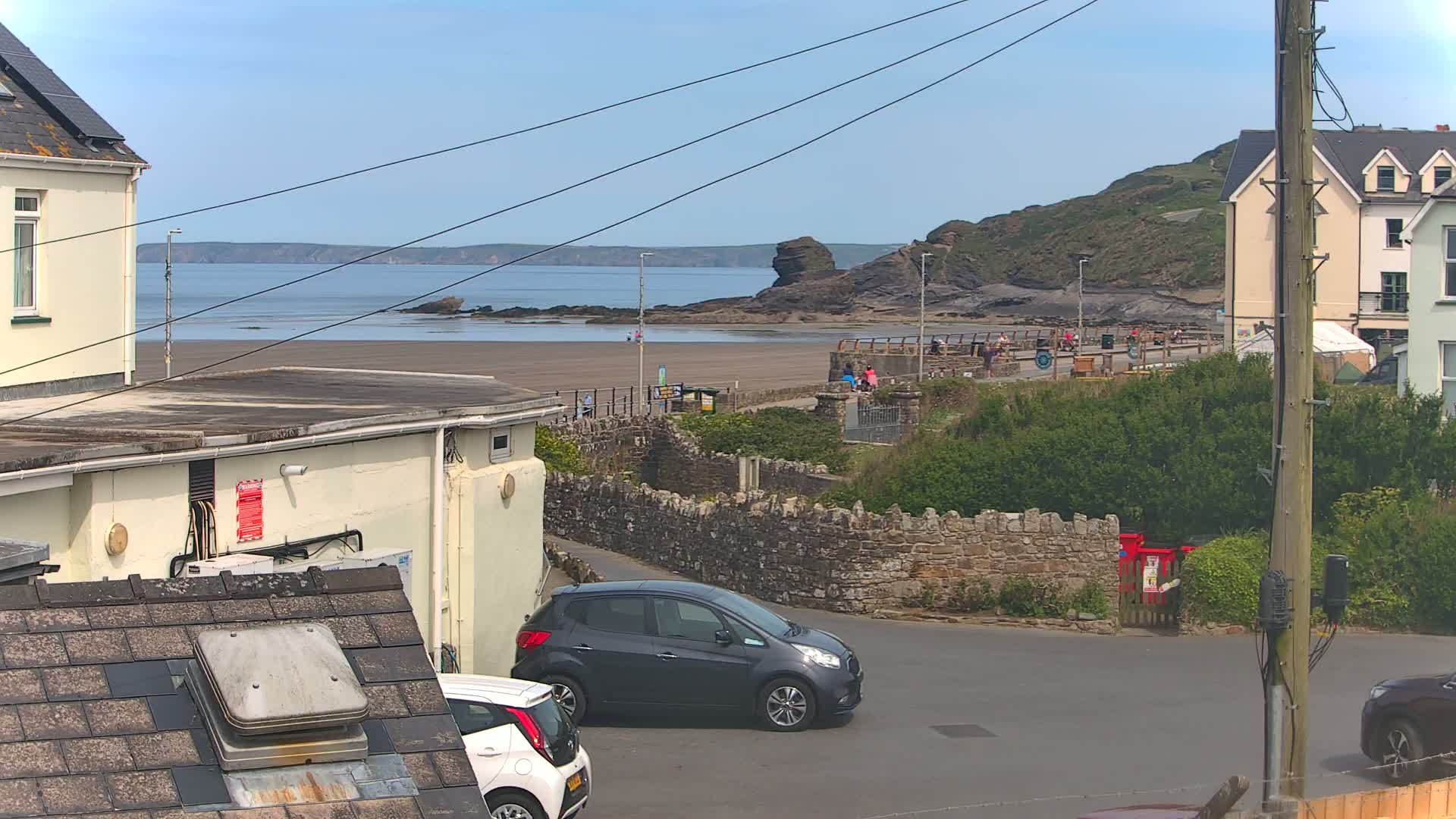 A sandy beach with calm ocean water stretches below a seaside town on a sunny day; two cars are parked near a stone wall separating a road from the beach.