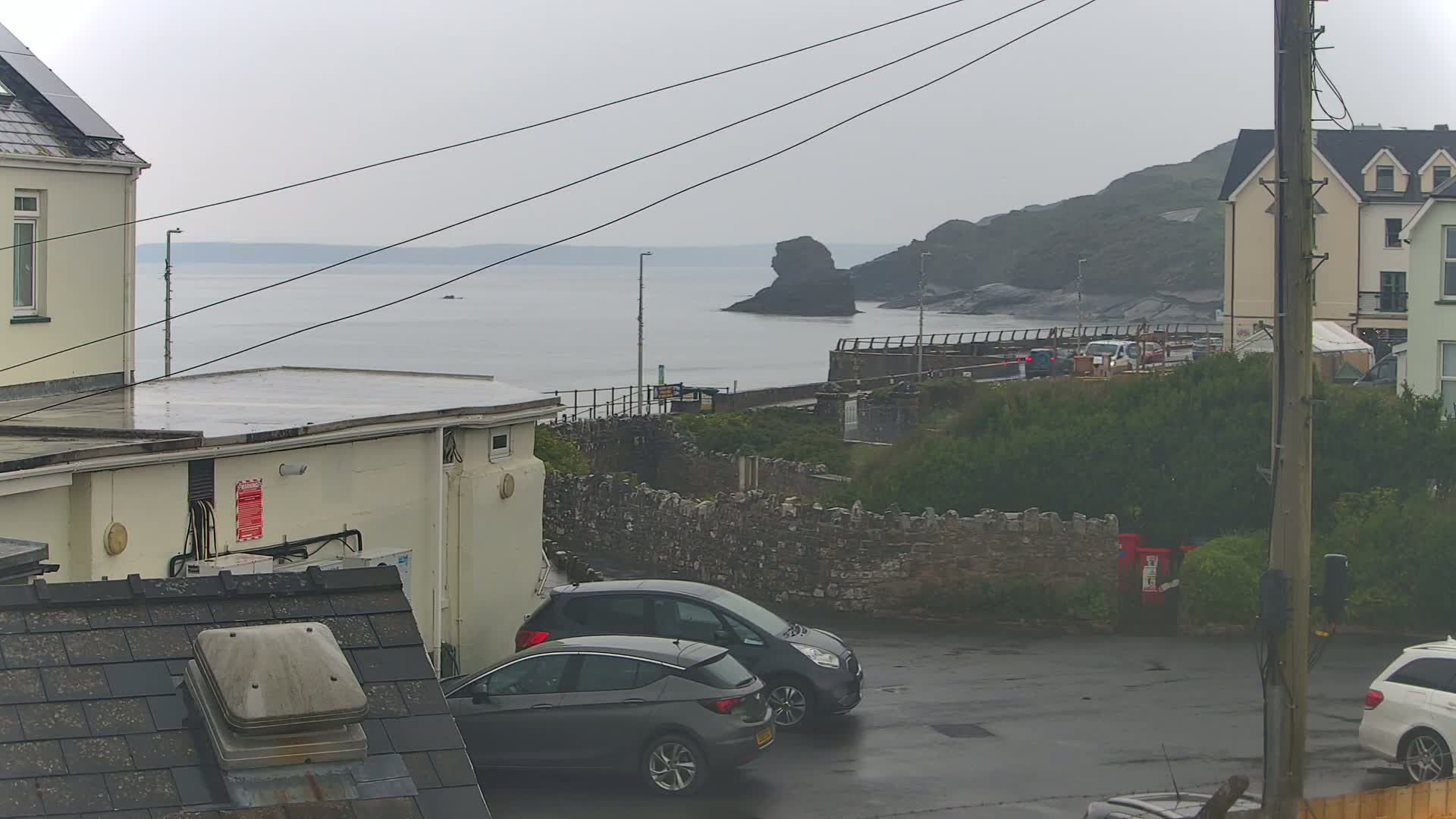 A wet, overcast coastal scene shows two cars parked in a lot next to a stone wall overlooking a calm sea with a rocky outcropping.