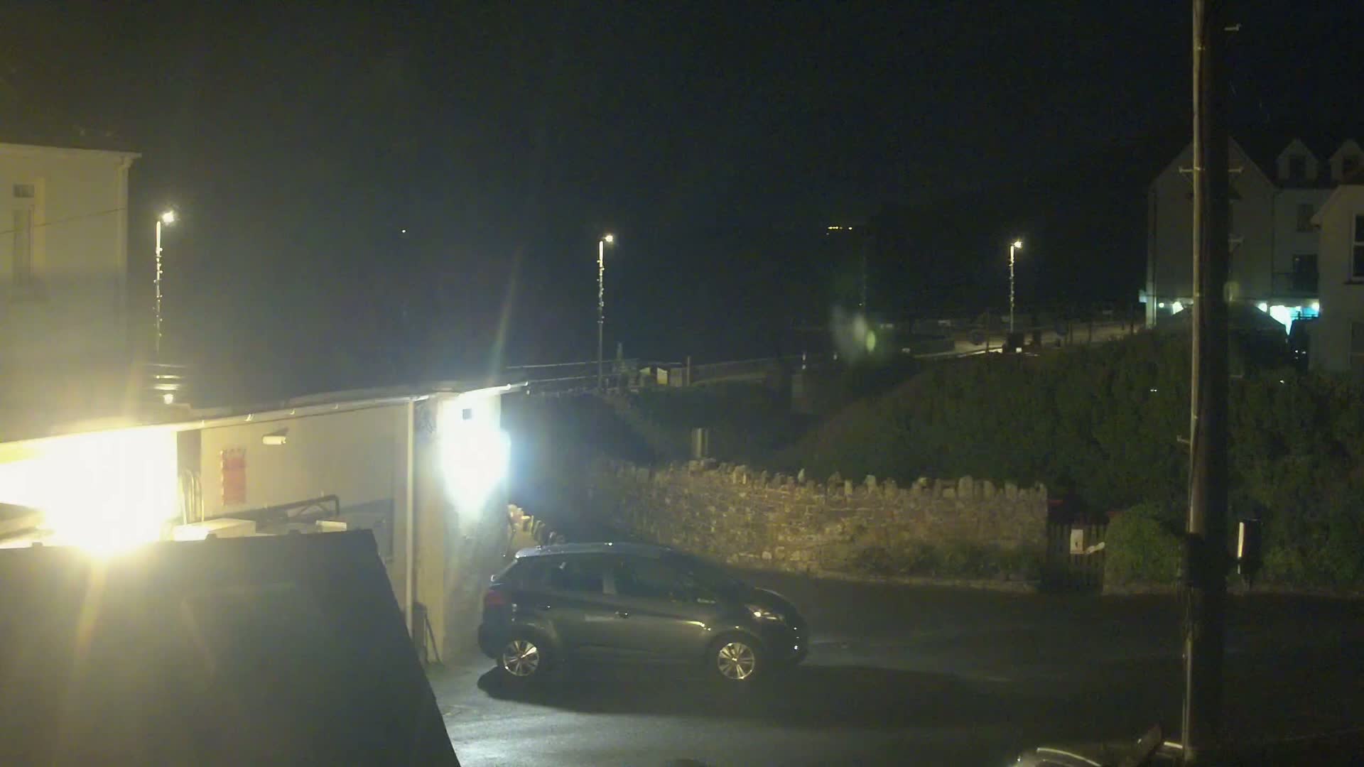 A dark-colored car is parked in a dimly lit area at night, near buildings and a low stone wall.