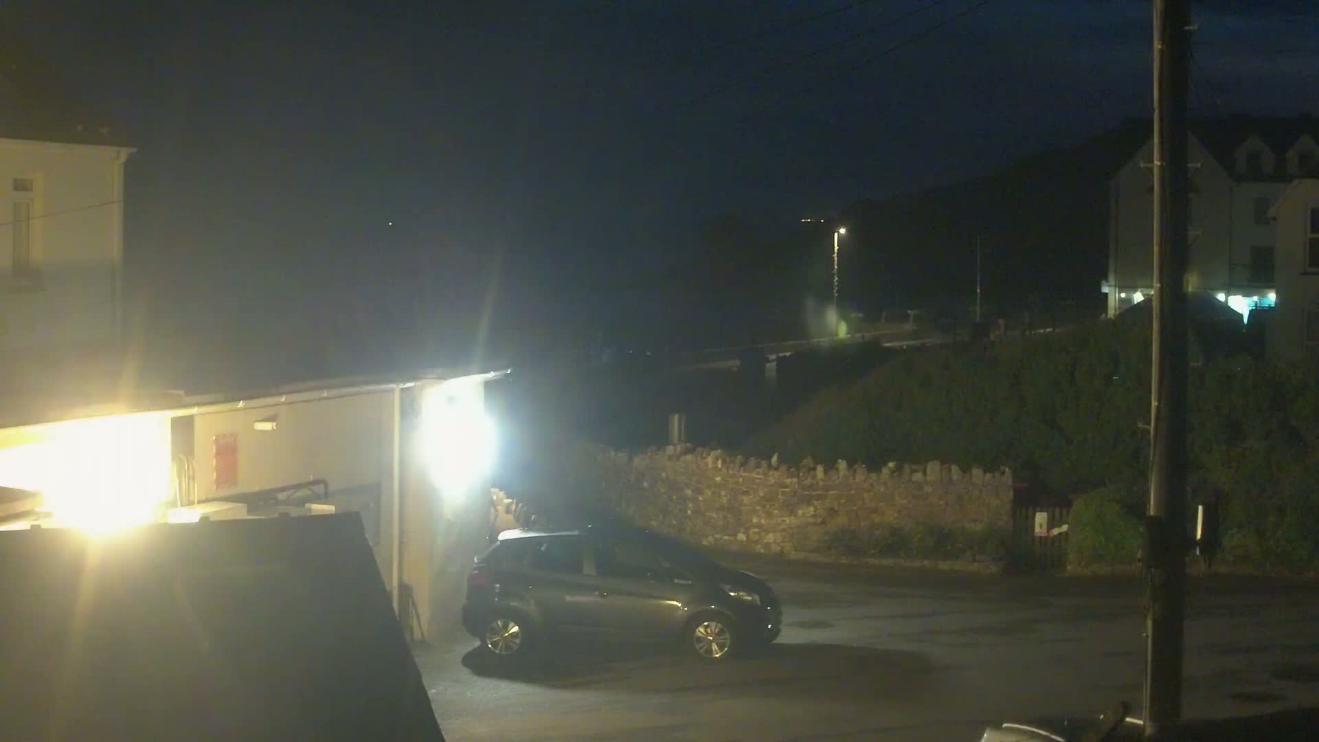 A dark-colored car is parked in a dimly lit area at night, next to a low stone wall and buildings under a dark, possibly overcast sky.