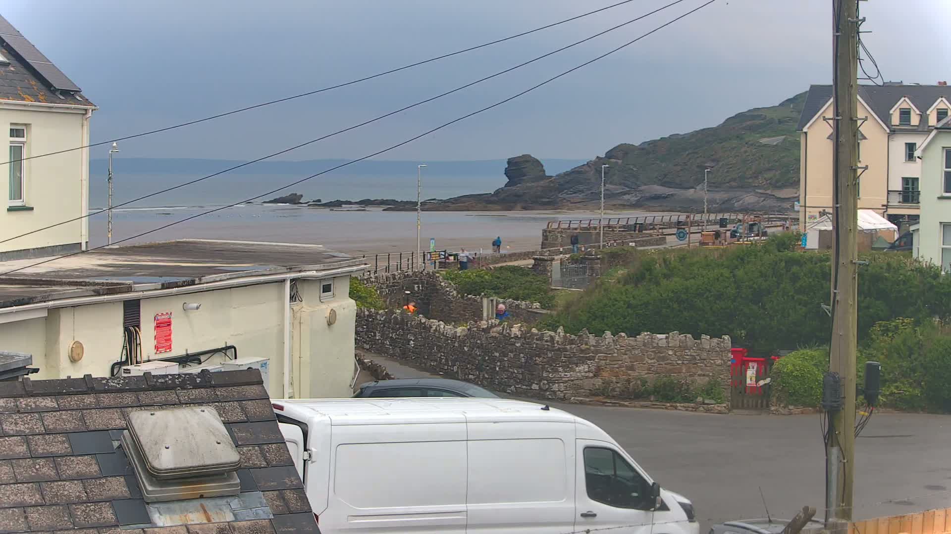A view of a coastal town on an overcast day, showing a sandy beach, rocky shoreline, buildings, and a white van in the foreground.