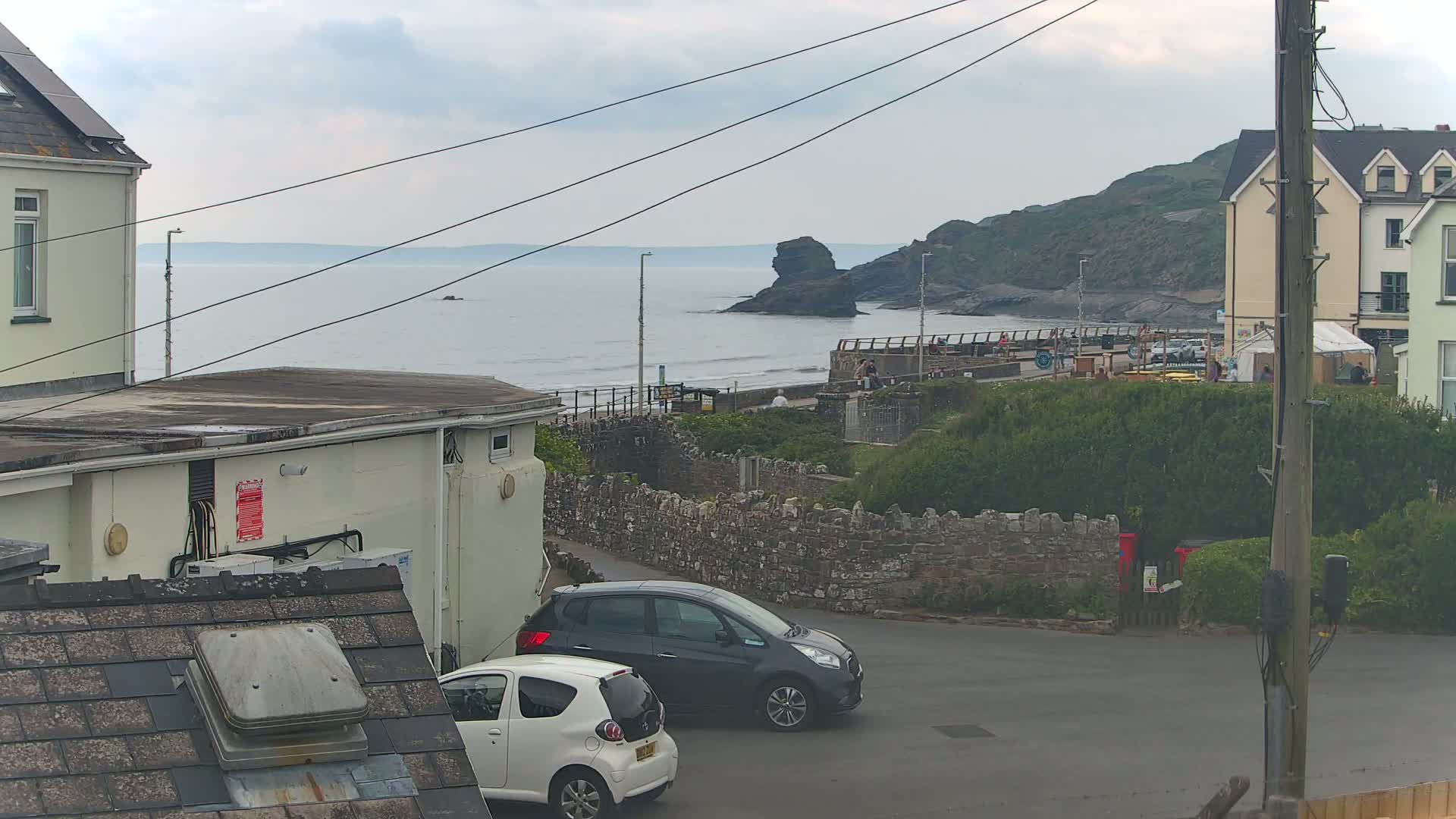 Overcast seaside view of a town with parked cars, buildings, and a rocky coastline with people visible on a walkway near the water.