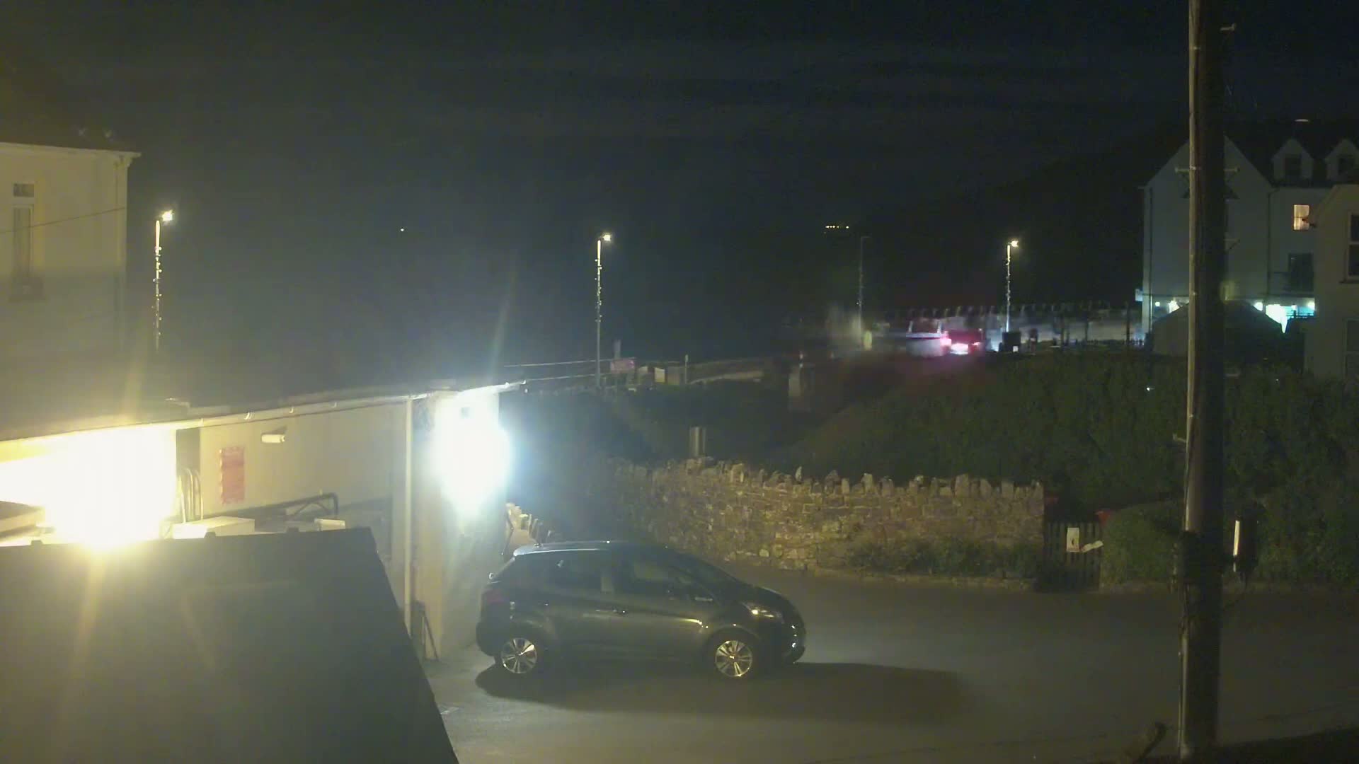 At night, a dark-colored car is parked in a lot near buildings and a low stone wall, with a few other vehicles visible in the distance under a dark sky.