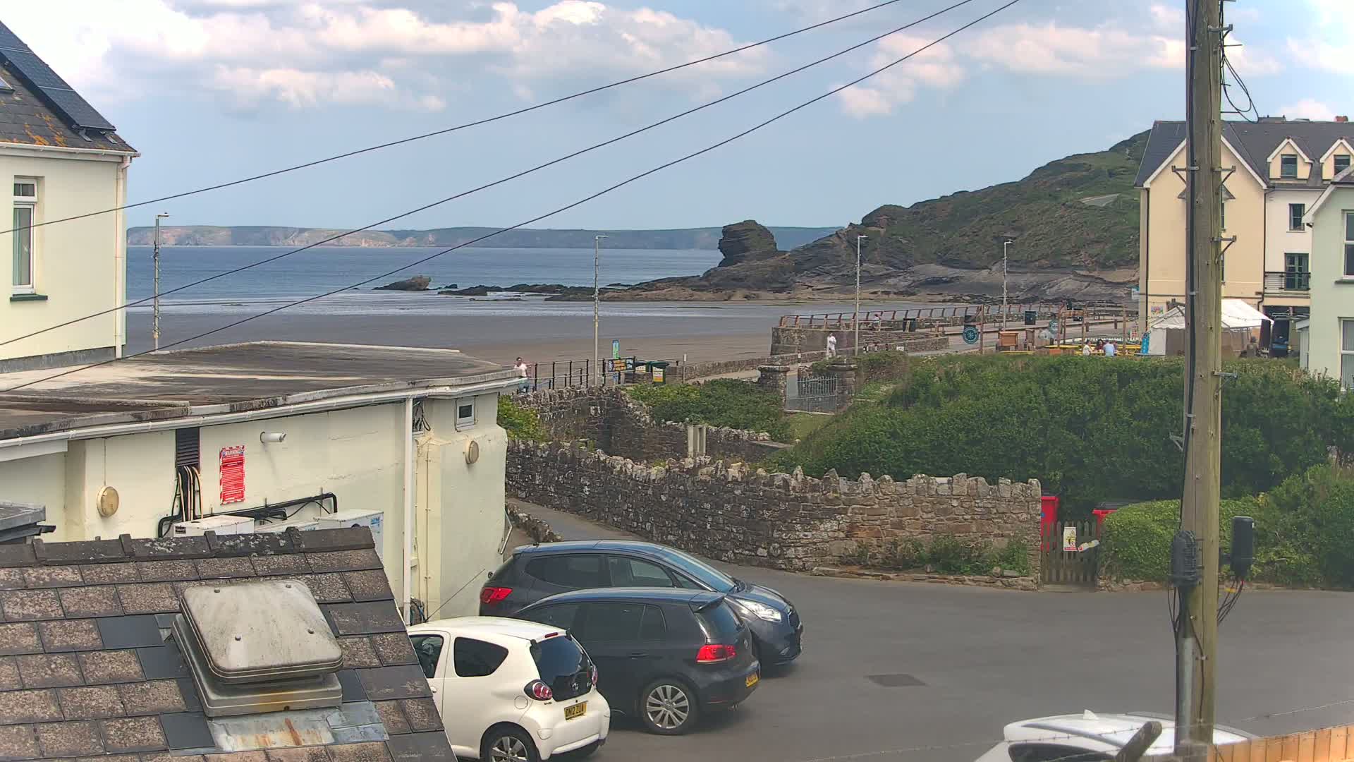A partly cloudy day overlooking a sandy beach with a rocky outcrop and calm sea, with several parked cars in the foreground and buildings visible on both sides of the street.