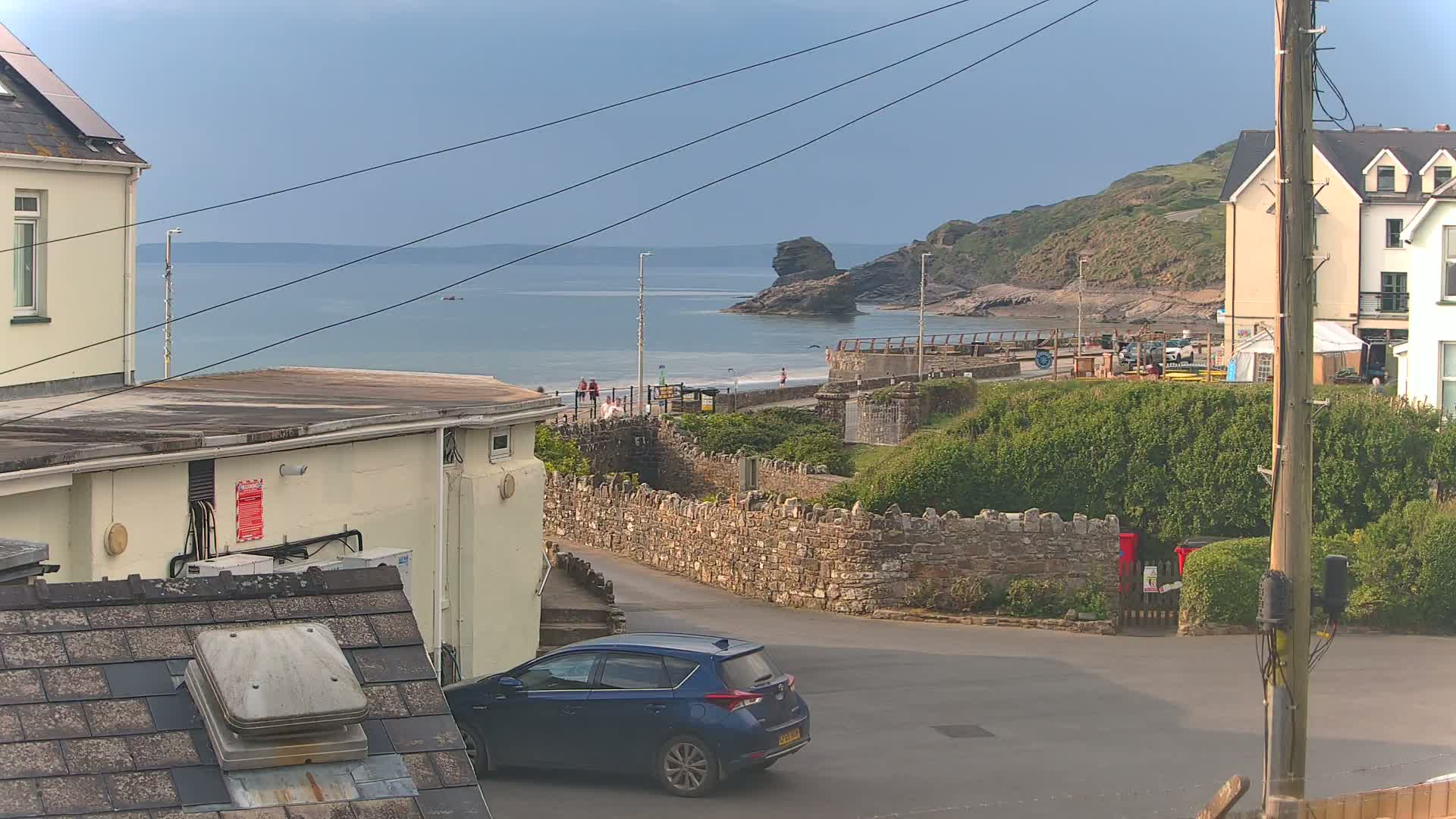 A blue car is parked near a stone wall overlooking a calm ocean, a beach with few people, and buildings on a partly cloudy day.