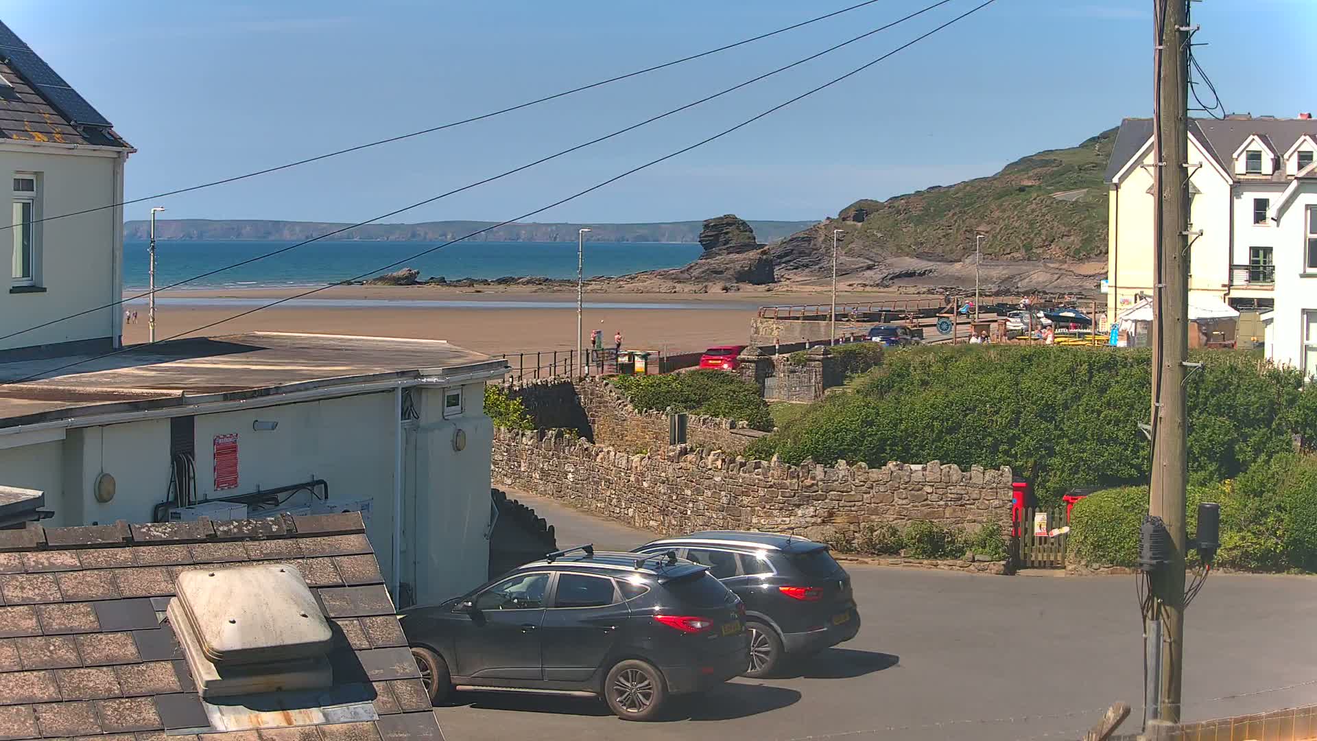 A sandy beach with calm ocean waters under a clear blue sky is viewed from a residential area with parked cars and buildings.