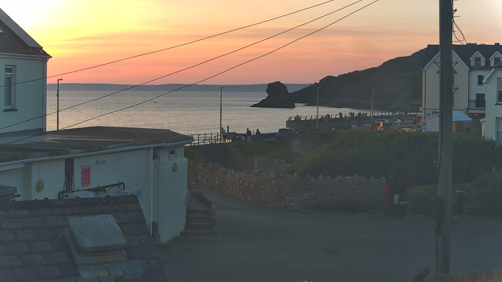 A seaside sunset view shows several buildings, a stone wall, people gathered on a coastal walkway, and a calm ocean with a rocky outcrop in the distance.