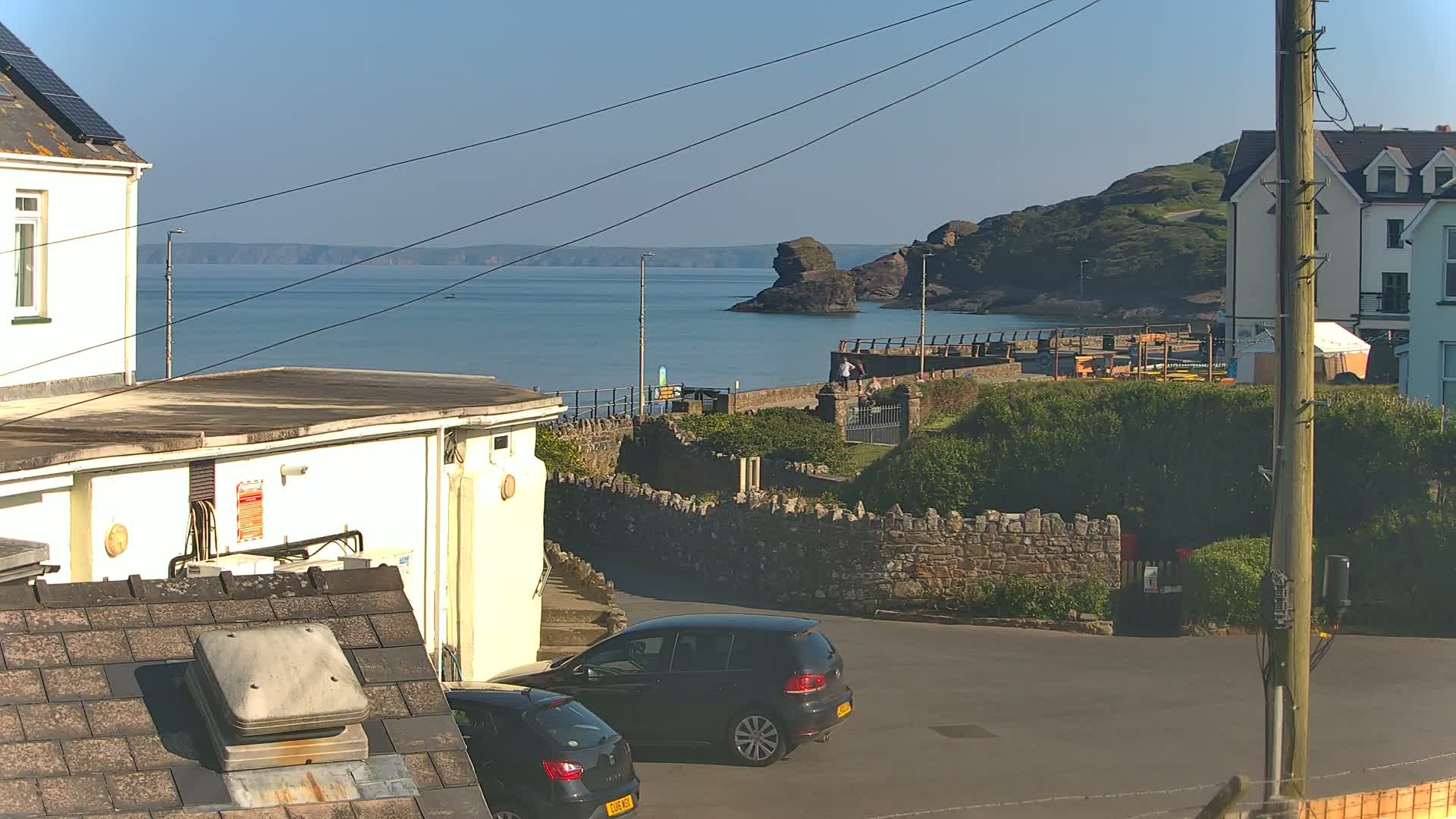 A coastal scene on a sunny day shows a calm sea with a rocky outcropping, a stone wall, several buildings, and two cars parked in a small lot.