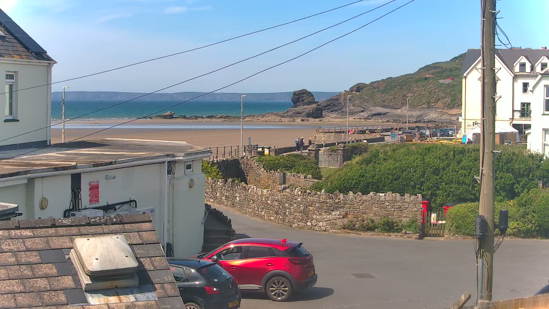 A sunny seaside scene is viewed from a rooftop, showing a sandy beach, calm ocean, rocky cliffs, buildings, and two cars parked on a street below.