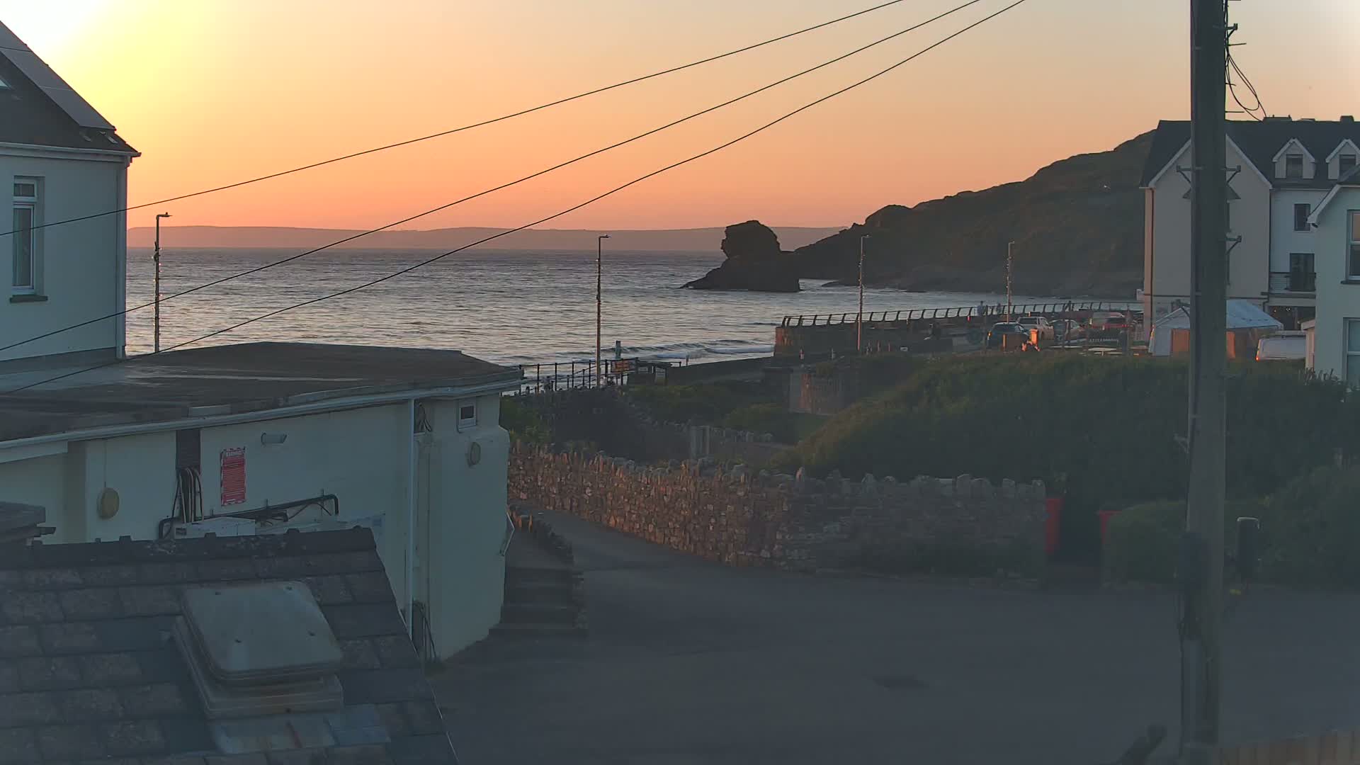 A coastal sunset view shows houses, a stone wall, and a calm sea with a rocky outcrop under a pinkish-orange sky.