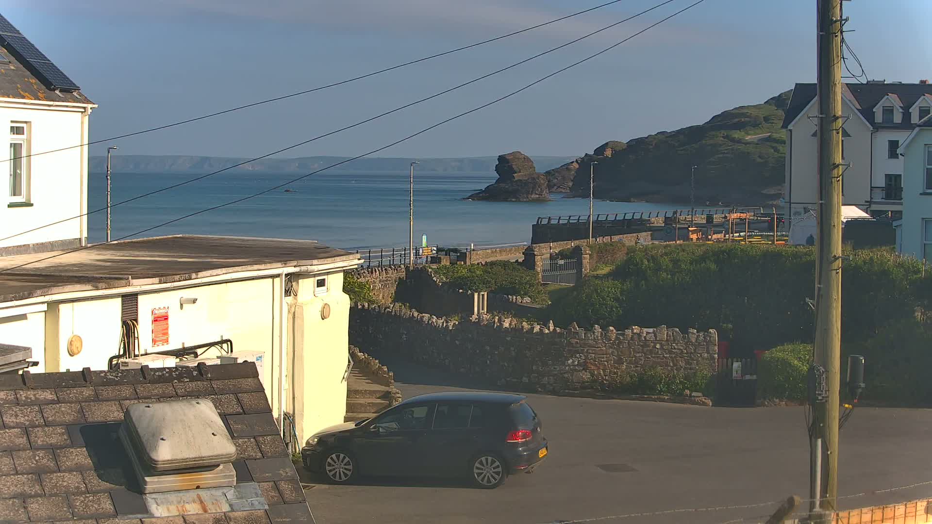 A dark-colored hatchback is parked in a seaside town on a sunny day, with a calm ocean and rocky coastline in the background.