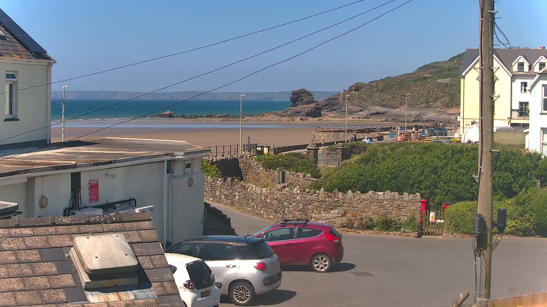 A sunny seaside view shows a sandy beach, calm ocean, rocky cliffs, buildings, and two parked cars.