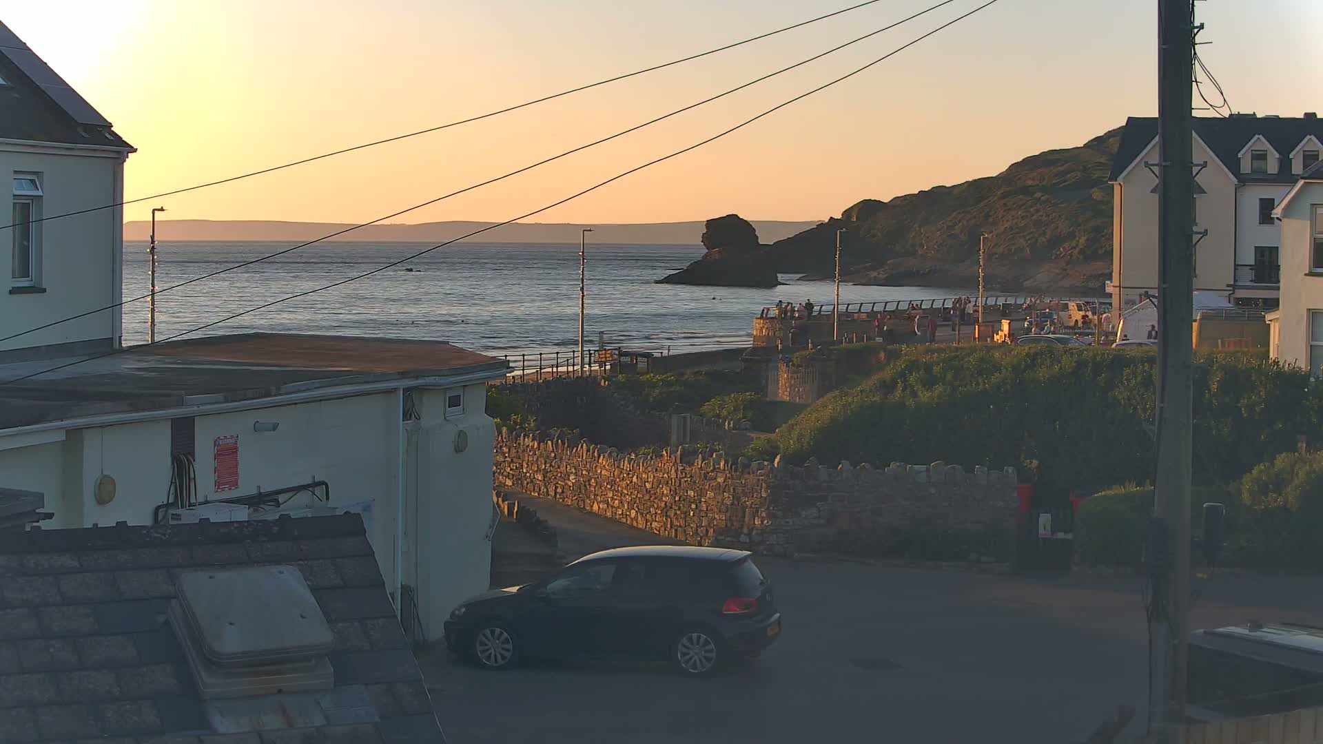 A dark-colored car is parked in a seaside town at sunset, with a calm ocean, a rocky coastline, and several buildings visible.