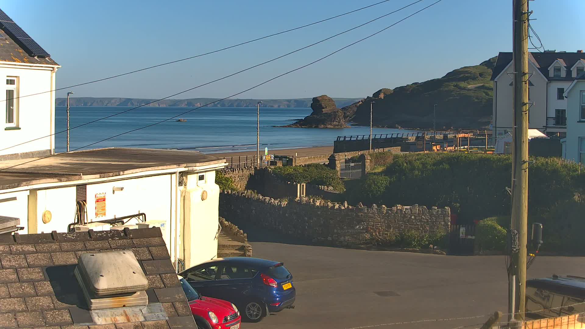 A sunny seaside view shows a sandy beach, calm ocean, distant cliffs, buildings, and two parked cars.
