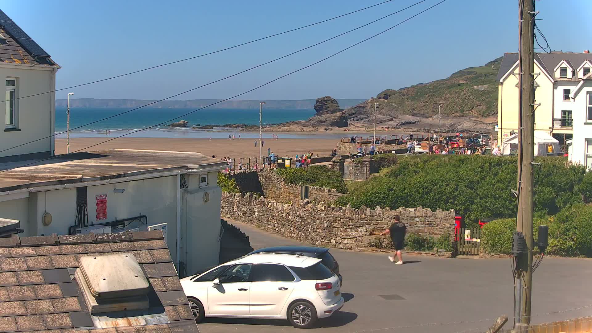 A sunny day at a beach with people on the sand and a few cars parked near buildings on a hillside.
