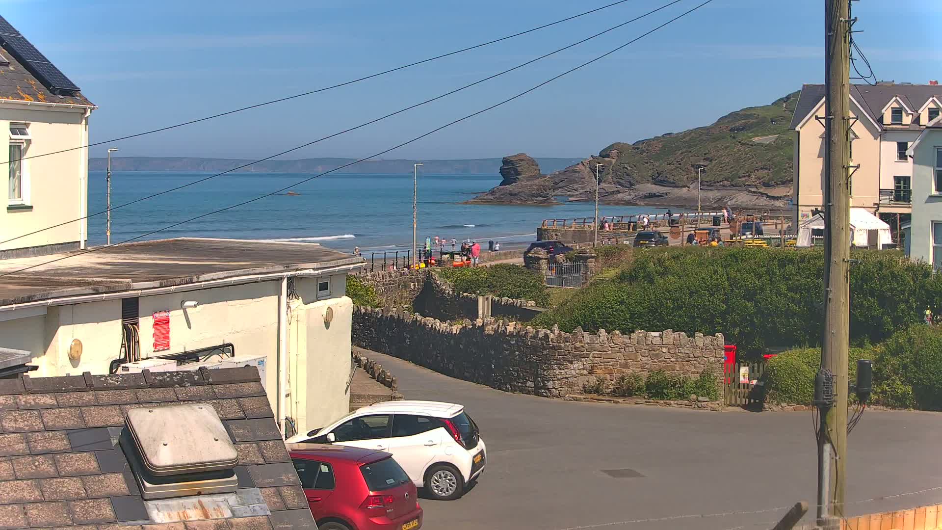 A sunny coastal scene shows a sandy beach with people, ocean waves, and buildings, with parked cars visible in the foreground.