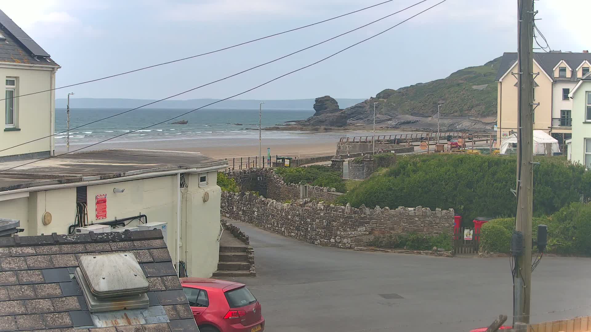 A view of a sandy beach with small waves under an overcast sky, seen from a residential street between buildings, with a red car parked nearby.