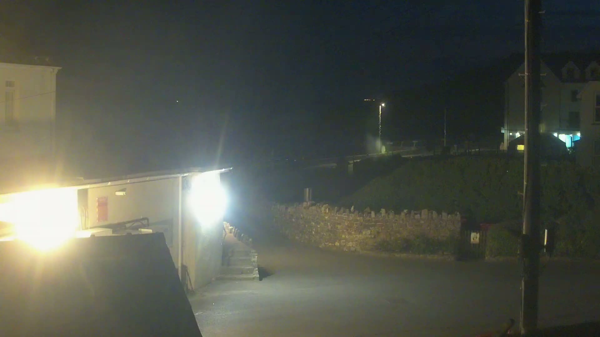 At night, brightly lit buildings are partially visible, along with a low stone wall and a dark, empty street.
