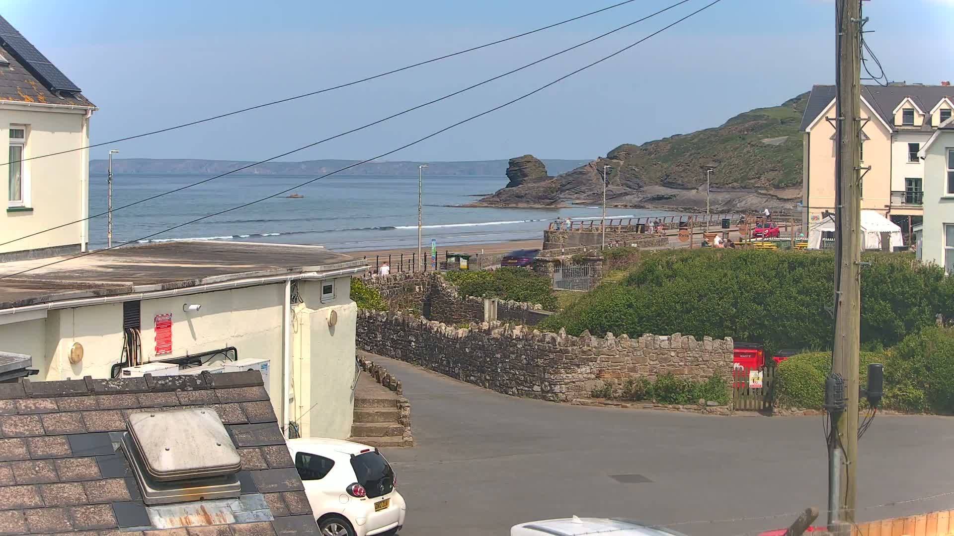 A sunny coastal scene shows a sandy beach with small waves, buildings, and a stone wall, with a small white car parked near a road.