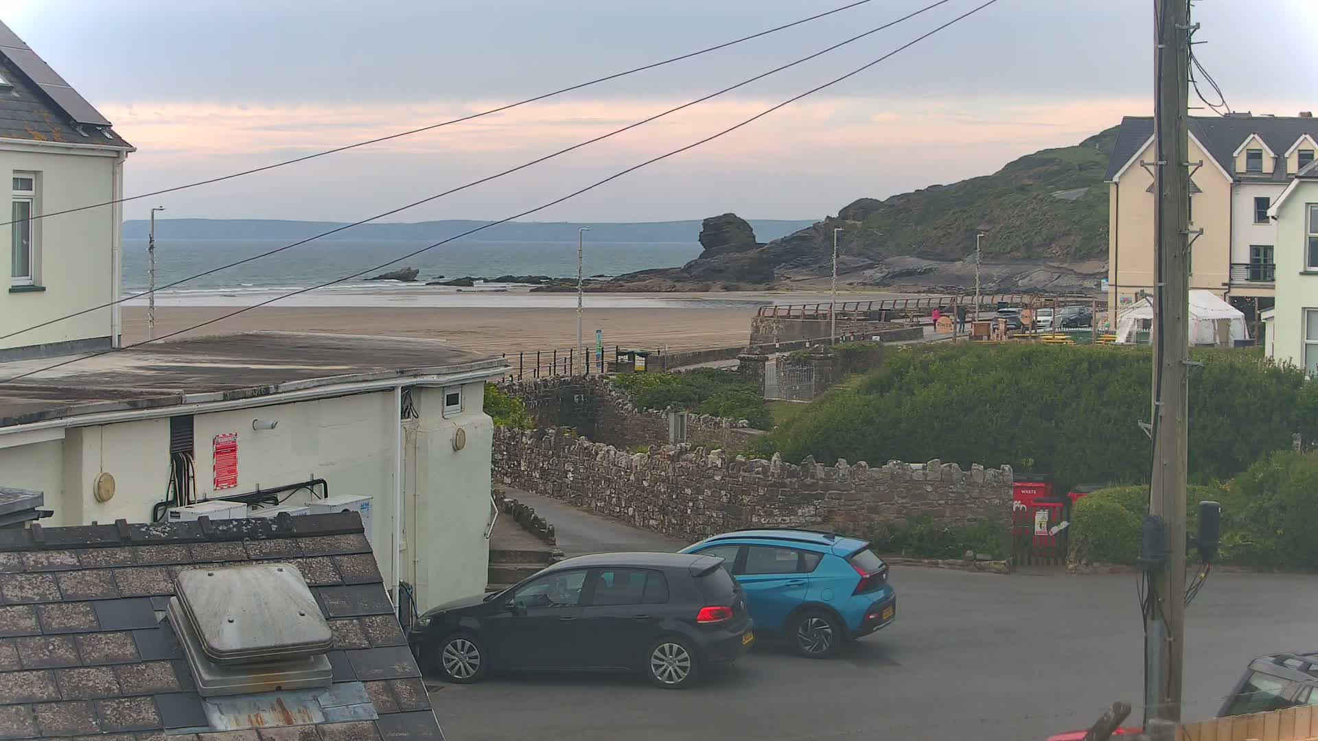 Overcast skies loom over a sandy beach and calm ocean, with two parked cars visible near buildings in the foreground.