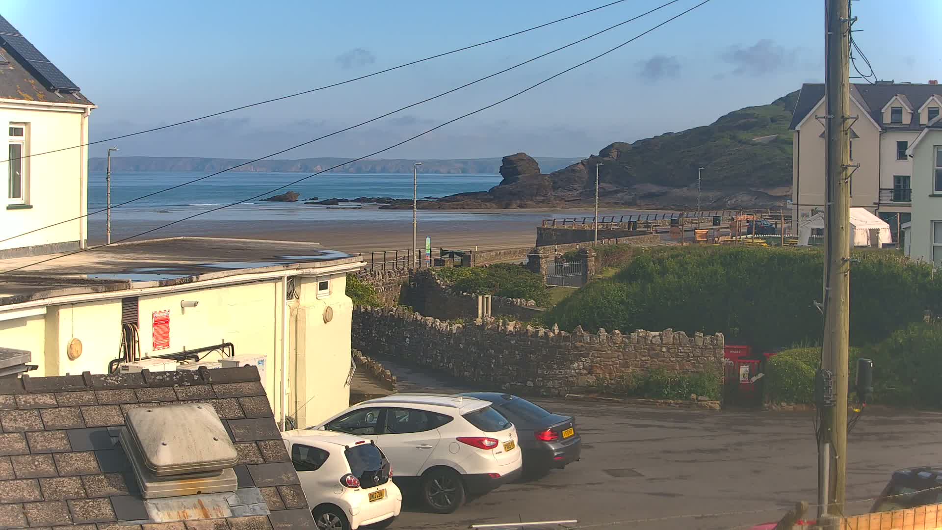 Two cars are parked in a lot overlooking a sandy beach and calm ocean under a mostly clear sky.