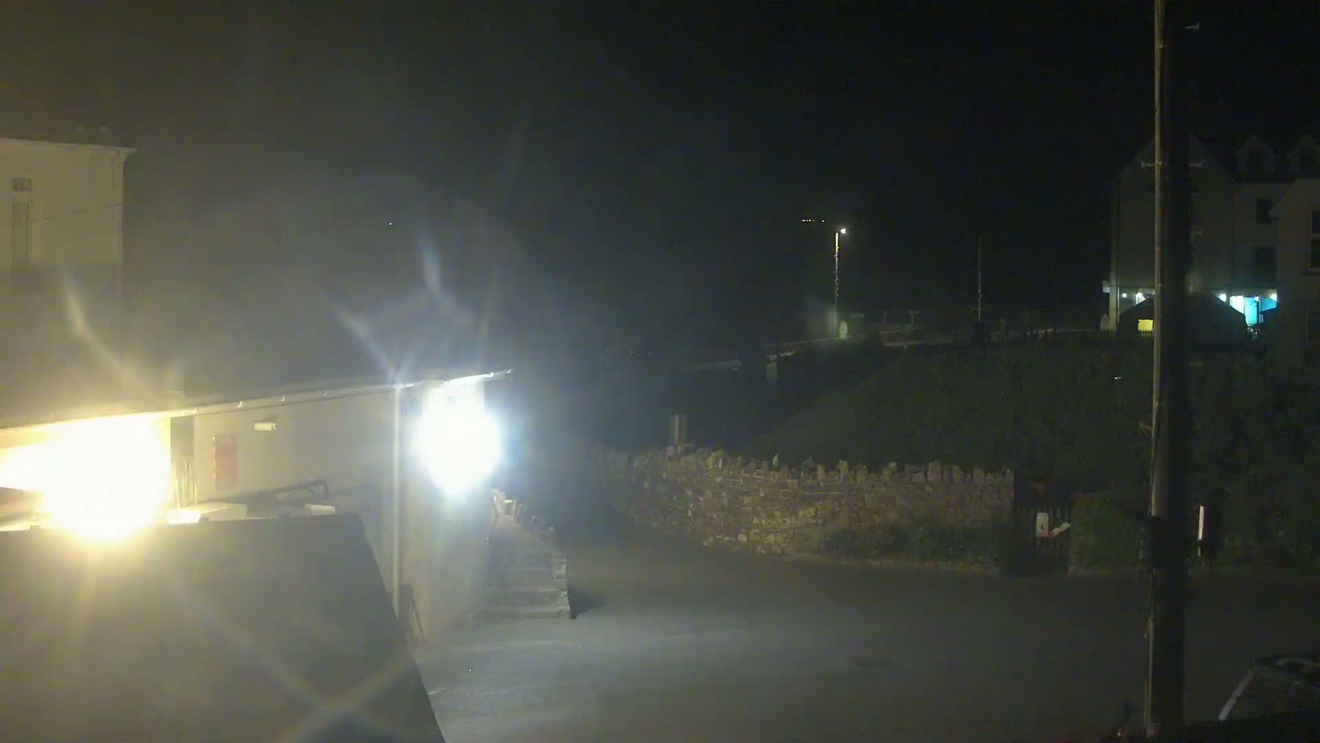 A nighttime view of a dimly lit street, showing buildings, a low stone wall, and bright exterior lights.