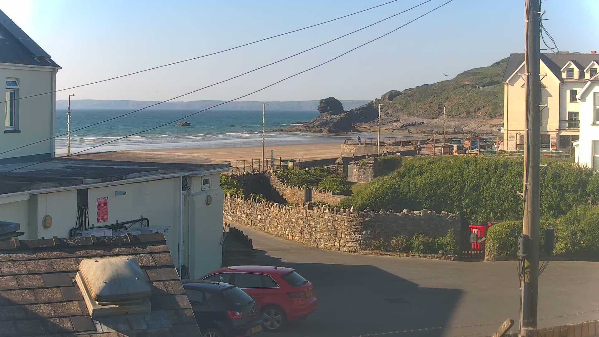 A sunny seaside town scene features a sandy beach with gentle waves, houses, and a red car parked near a stone wall.