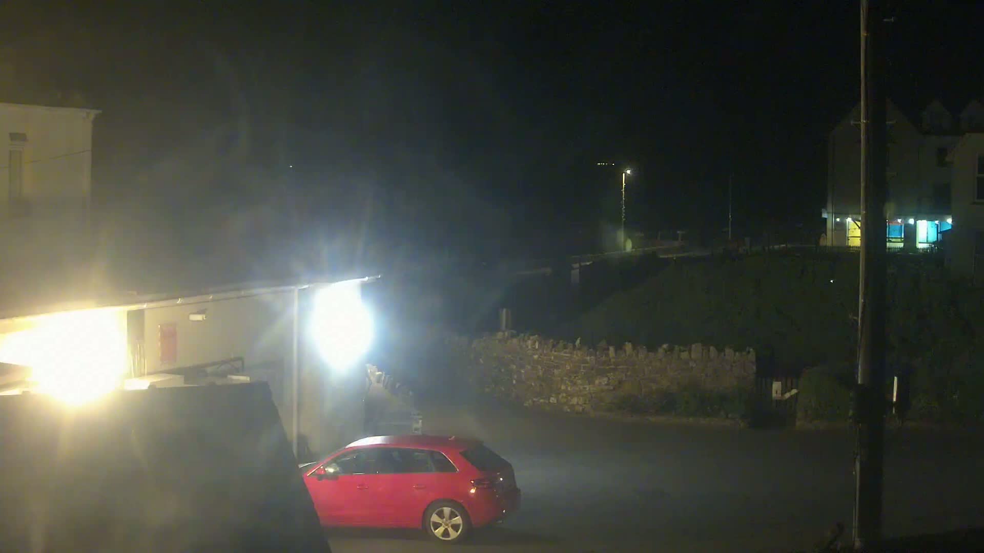 A red car is parked in a dimly lit area at night, near buildings and a low stone wall under hazy conditions.