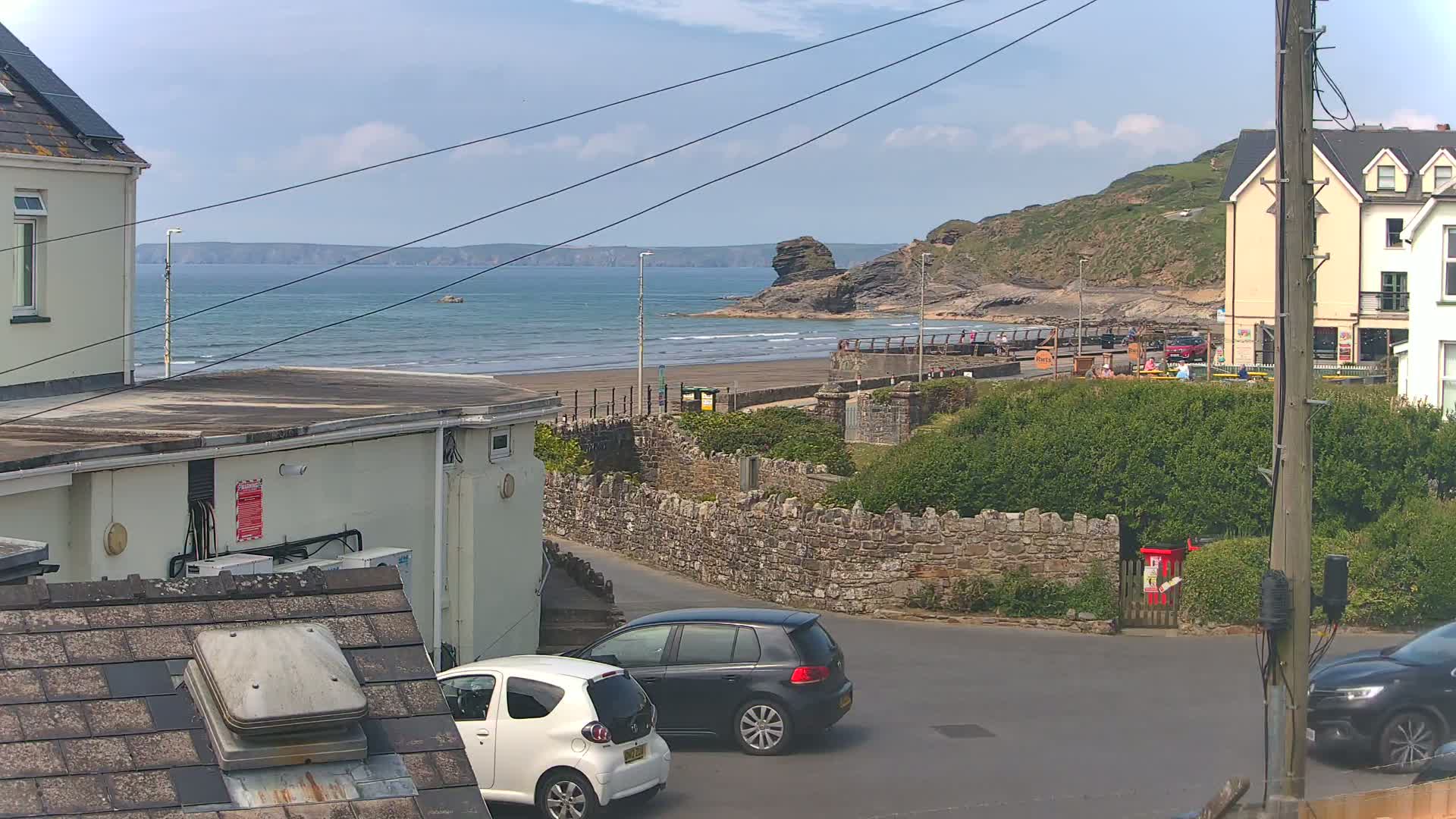 A seaside town scene on a partly sunny day shows a sandy beach with gentle waves, a rocky outcrop, buildings, parked cars, and a low stone wall.