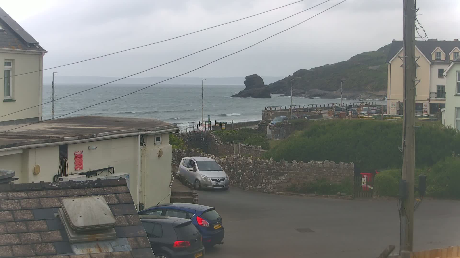 A view of a coastal town on an overcast day, showing a choppy sea, parked cars, buildings, and a stone wall.