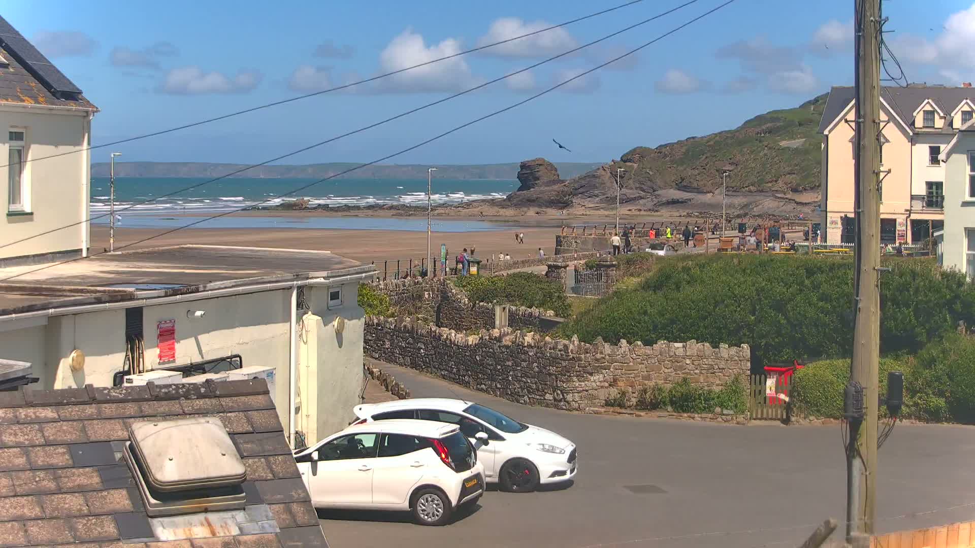Two white cars are parked near a stone wall overlooking a sandy beach with people walking along the shore under a mostly sunny sky with a few clouds.