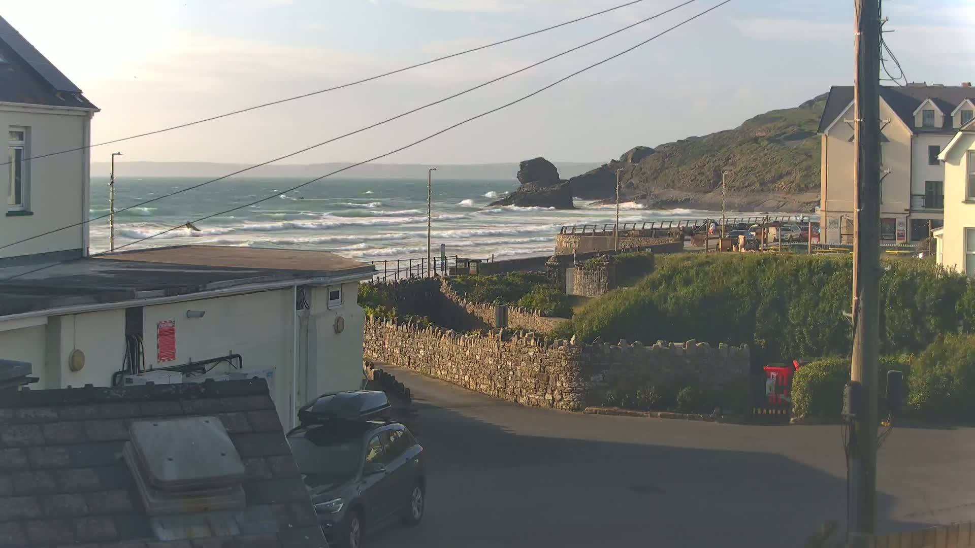 A view of a rocky coastline with moderate waves under a partly cloudy sky, seen from a residential area with houses and a car parked on a street.