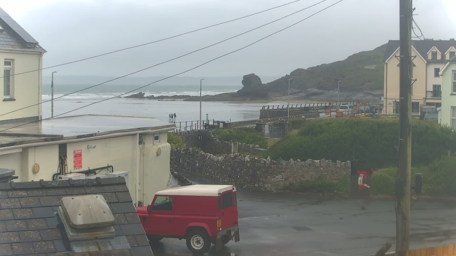 A red Land Rover is parked in a lot overlooking a gray, overcast beach with small waves and a few people walking near the water's edge.