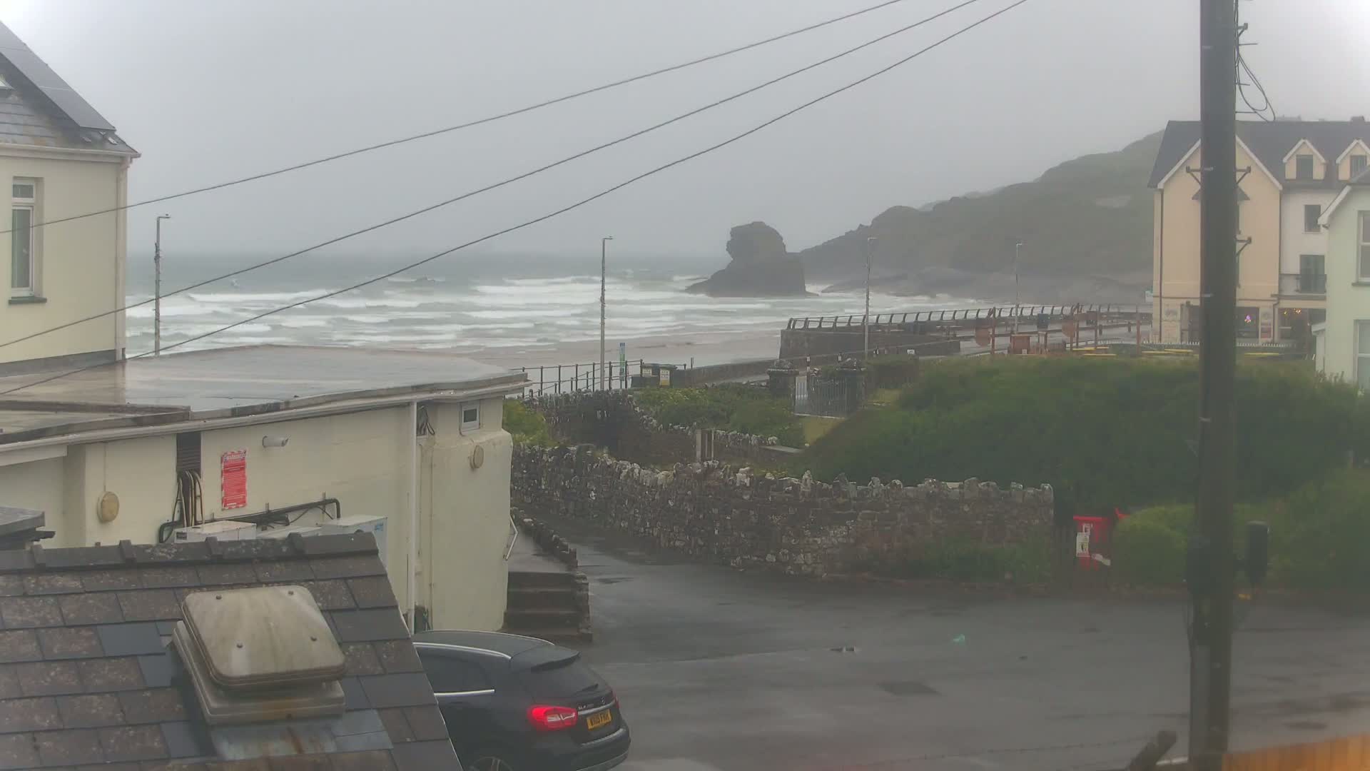 A rainy, overcast day reveals a coastal scene with choppy waves, a rocky outcrop in the distance, houses, a stone wall, and a dark-colored car parked near a building.