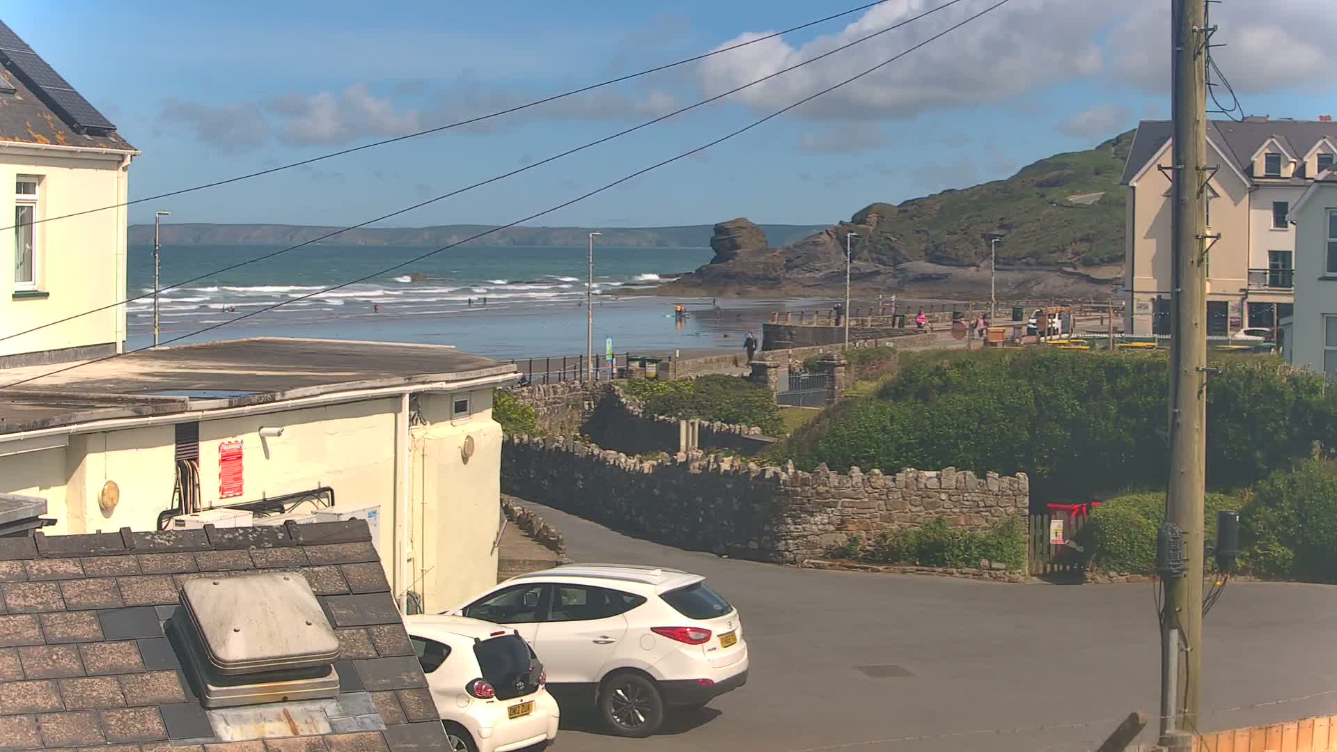 A sunny day at a beach with small waves, viewed from a residential area showing parked cars and buildings, with a rocky cliff in the background.