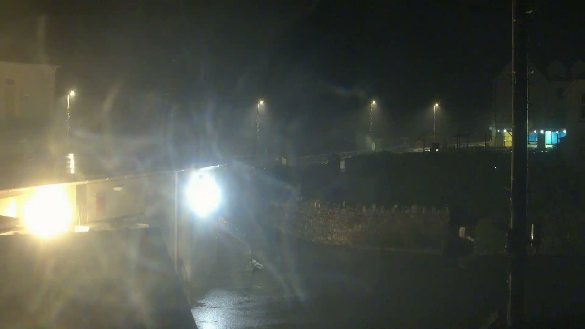 A nighttime, rainy scene shows a wet road and buildings illuminated by streetlights and headlights.