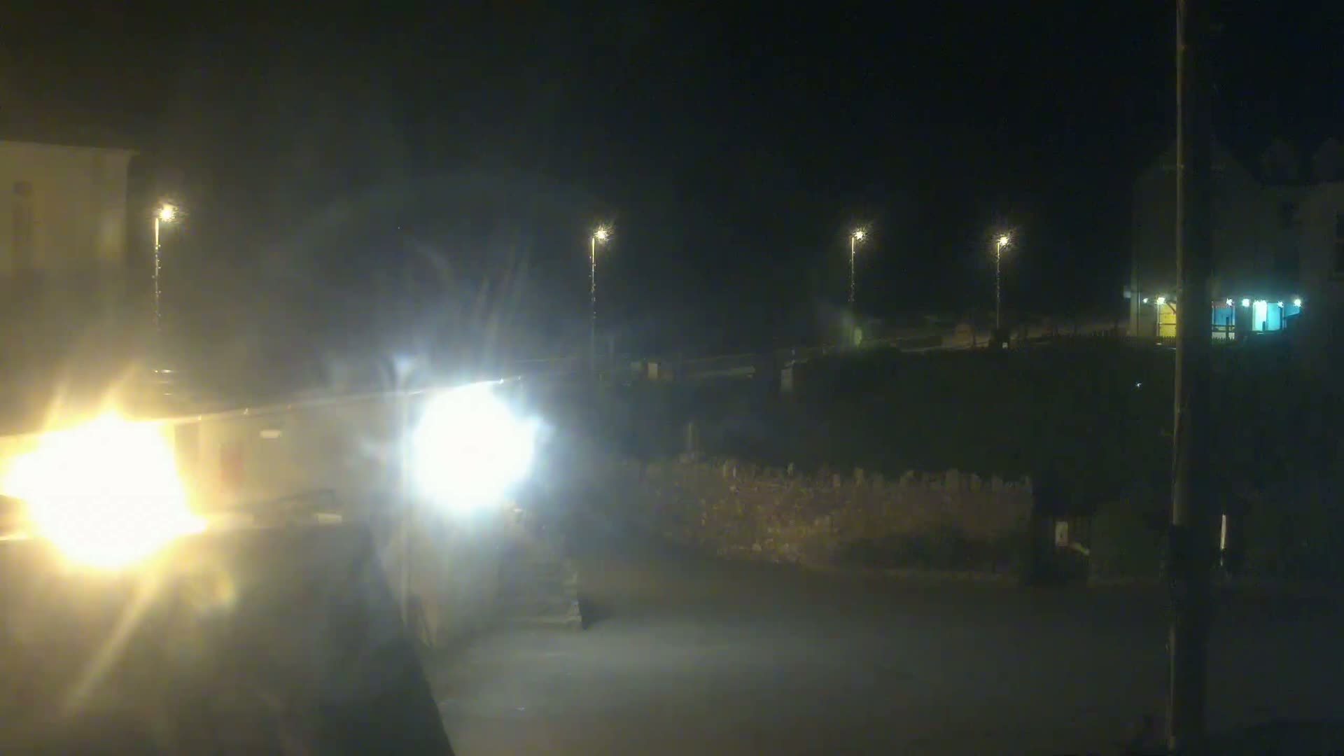 At night, a vehicle's headlights illuminate a dark street with a low stone wall and several streetlights in the background.