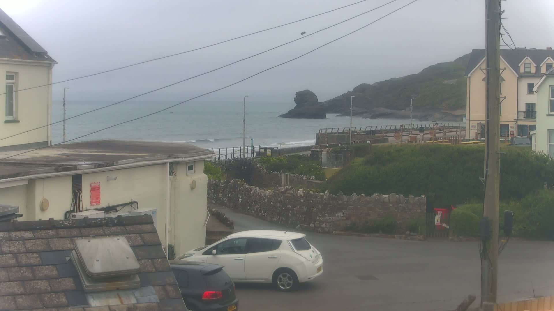 A grey, overcast day shows a coastal scene with a white electric car parked near buildings, a stone wall, and a calm sea with a rocky outcrop.