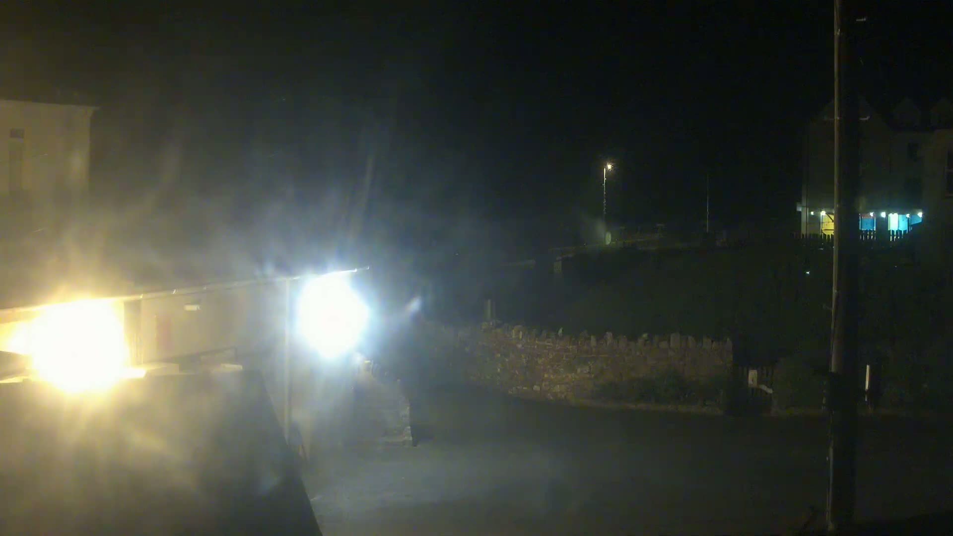 At night, in foggy conditions, a vehicle's headlights illuminate a street and nearby buildings.