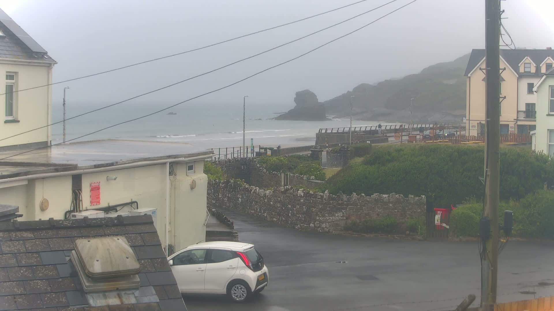 A misty seaside town scene shows a white car parked near buildings, a low stone wall, and a beach with a rocky outcrop in the distance.