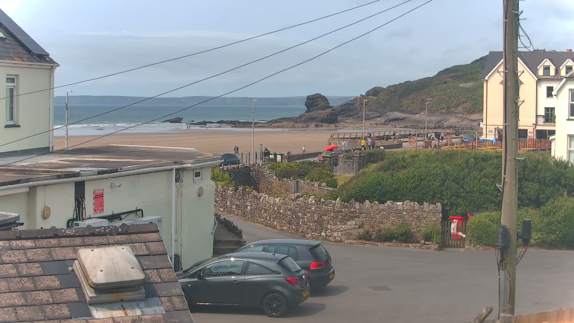A sandy beach with a few people on it is visible beyond a stone wall and parked cars, under a mostly cloudy sky.