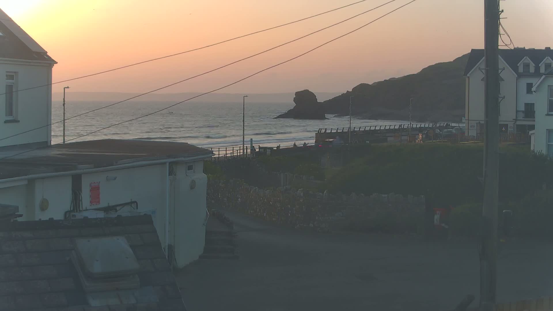 A sunset over a calm ocean viewed from between two buildings, with a rocky outcrop visible in the distance.