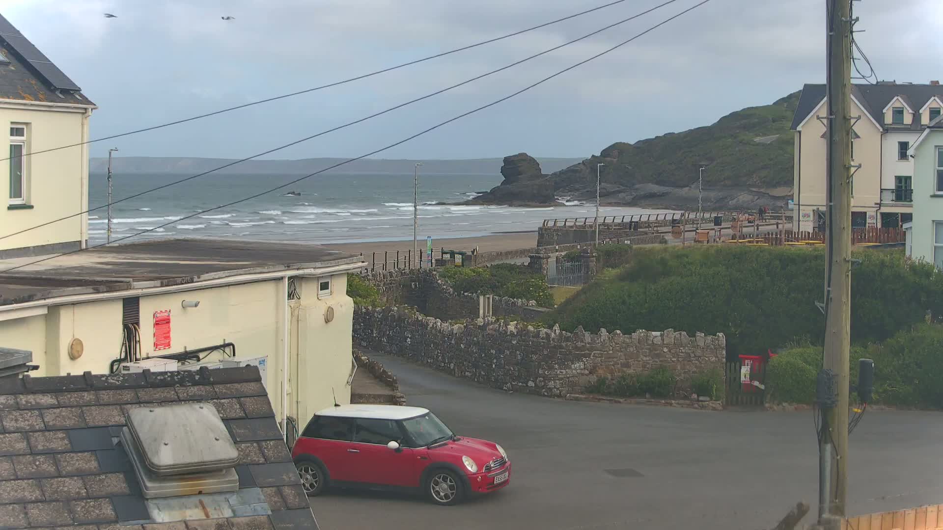 A red Mini Cooper is parked near a stone wall overlooking a beach with moderate waves under a cloudy sky.