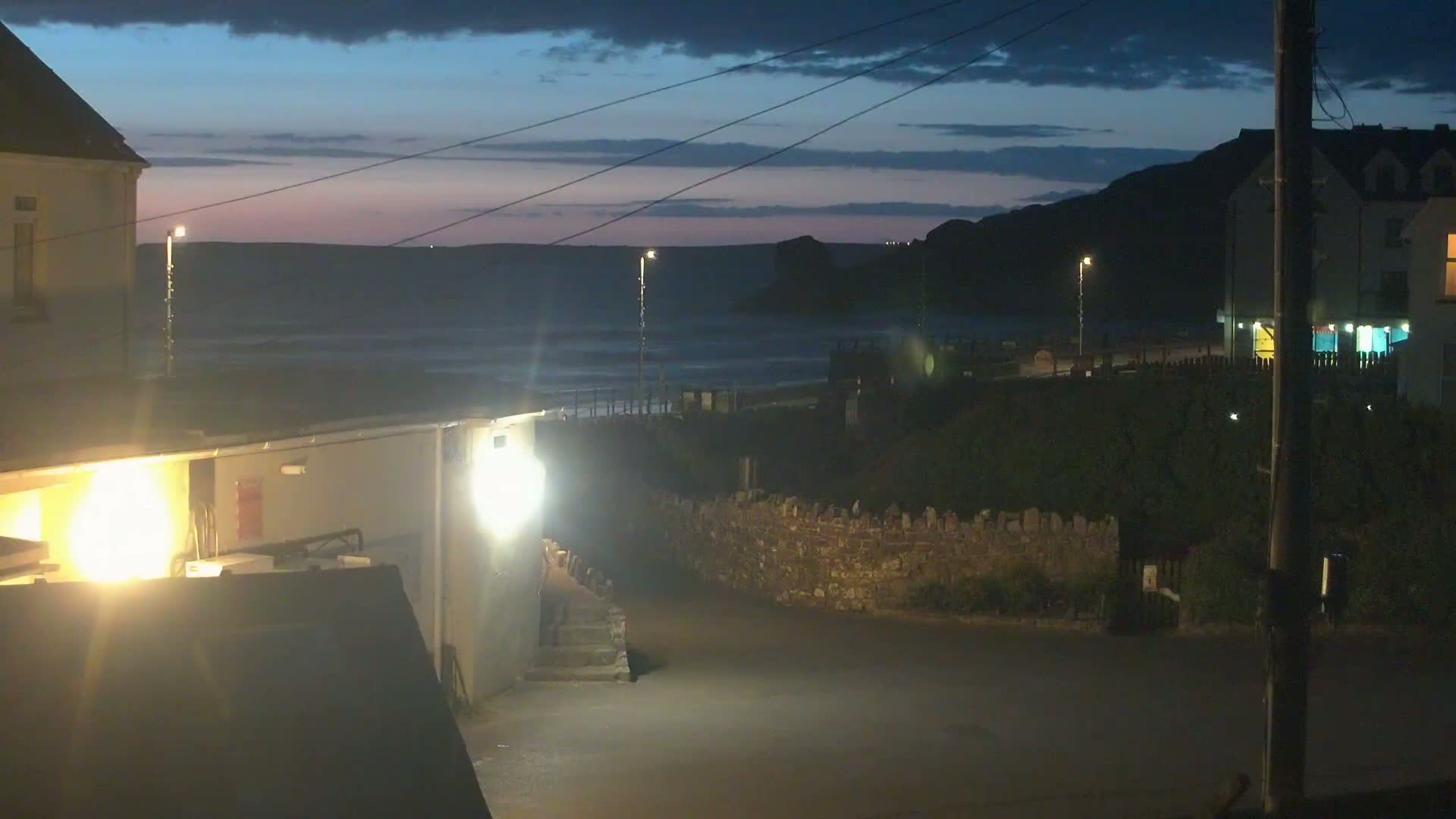 A nighttime coastal scene shows a dark ocean with waves under a dusky sky, buildings with lights illuminating a small paved area next to a low stone wall.