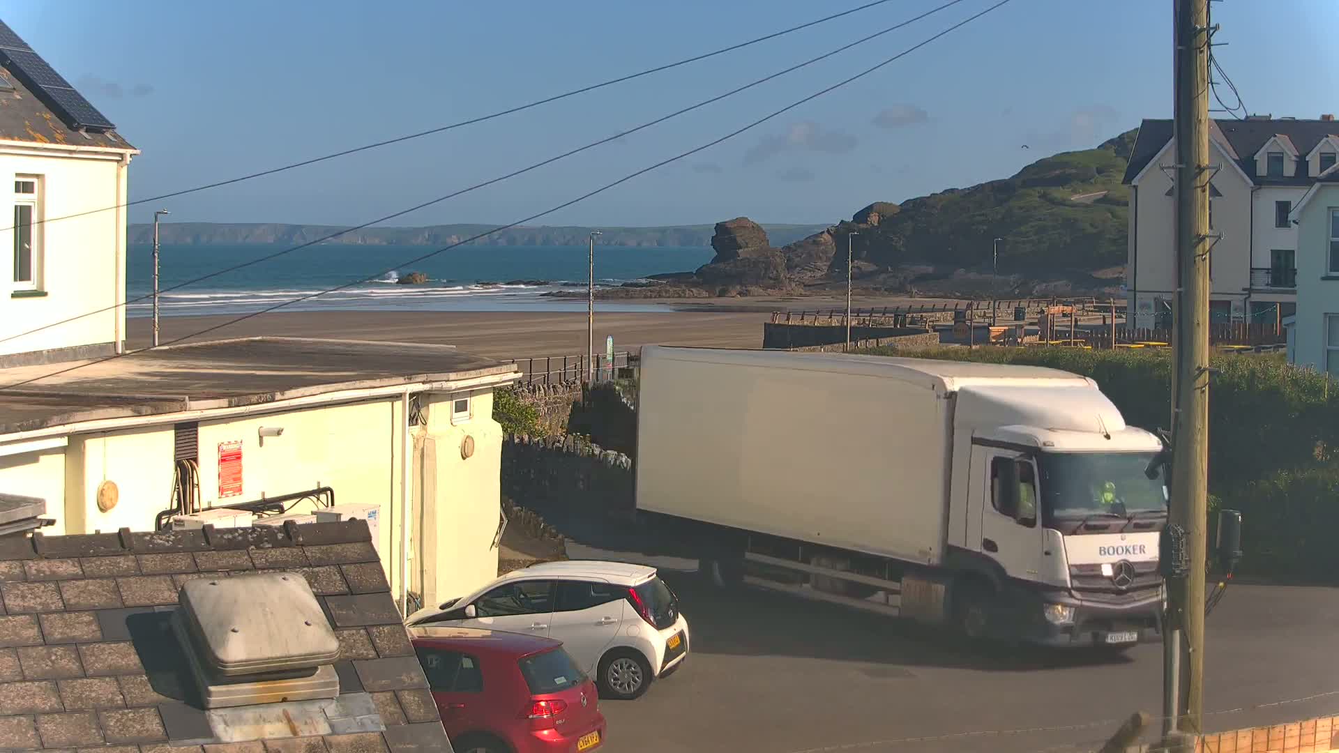 A white delivery truck is parked on a road near a sandy beach on a sunny day, with buildings and cars nearby.
