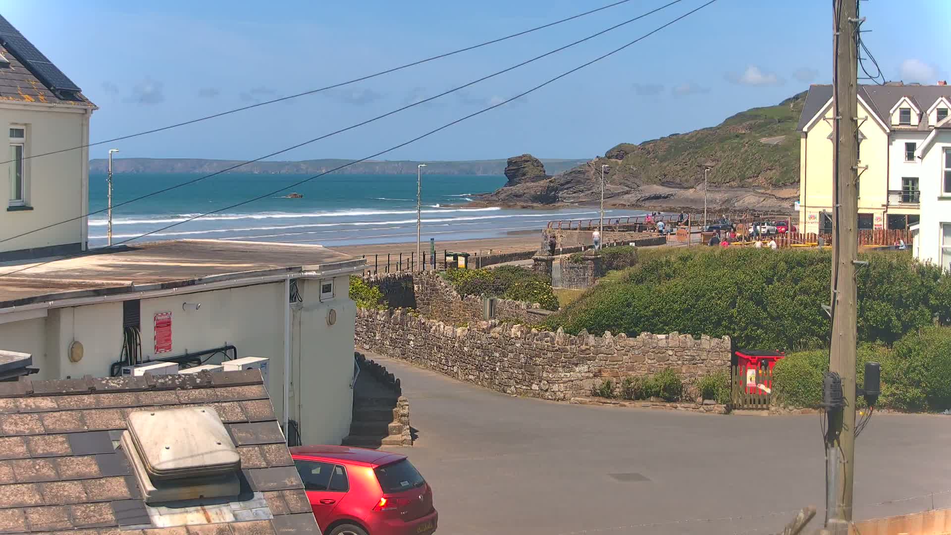 A sunny seaside town scene shows a sandy beach with gentle waves, a rocky outcrop, buildings, a red car, and a stone wall.